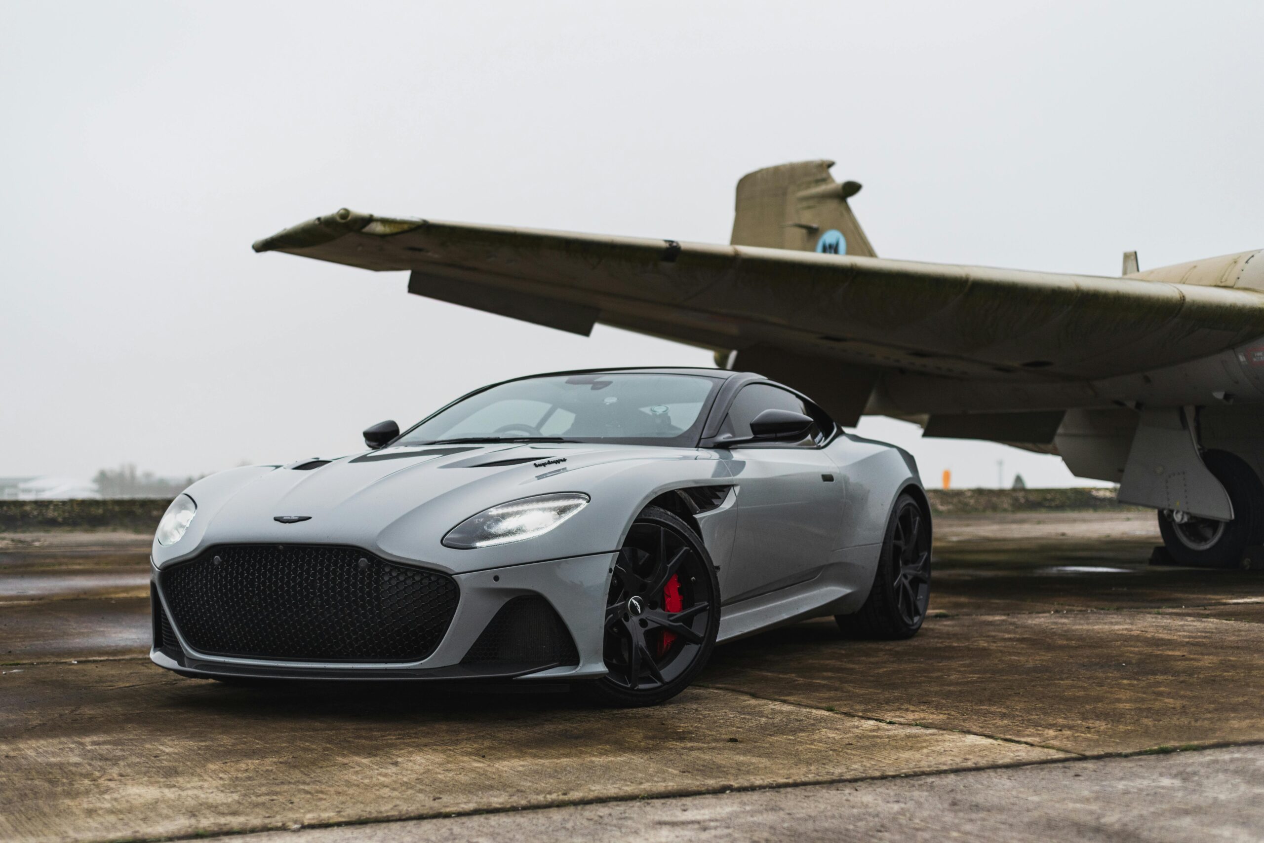 Sleek grey luxury car parked beneath an old aircraft on a foggy day.